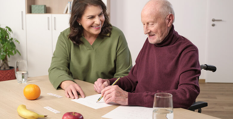 Senior schreibt eine Übung mit Unterstützung einer Betreuerin am Tisch