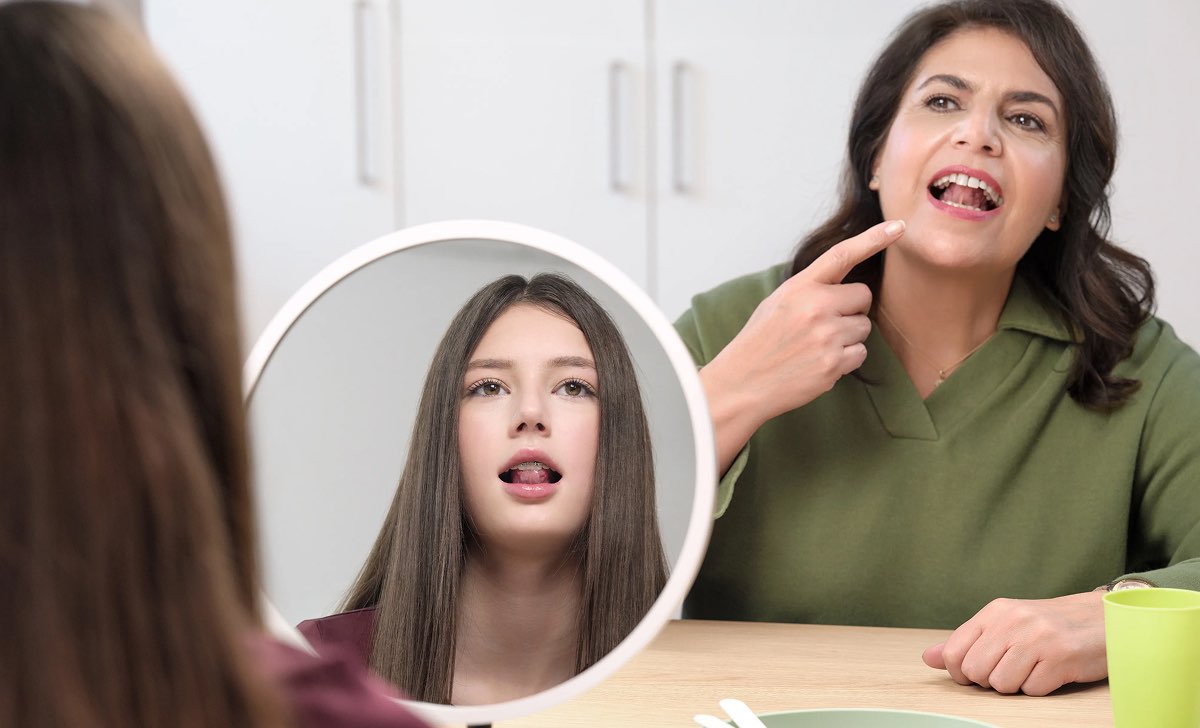 Therapeutin zeigt einem Mädchen vor dem Spiegel eine Mund- und Zungenübung in der Sprechtherapie
